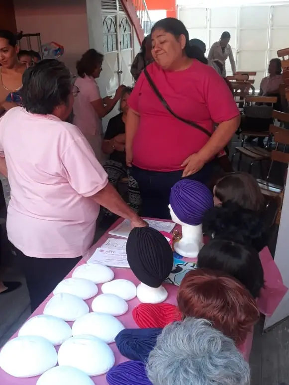 Mesa rosa con prótesis mamarias externas, turbantes y pelucas en una entrega de Grupo RETO Guadalajara, con mujeres alrededor